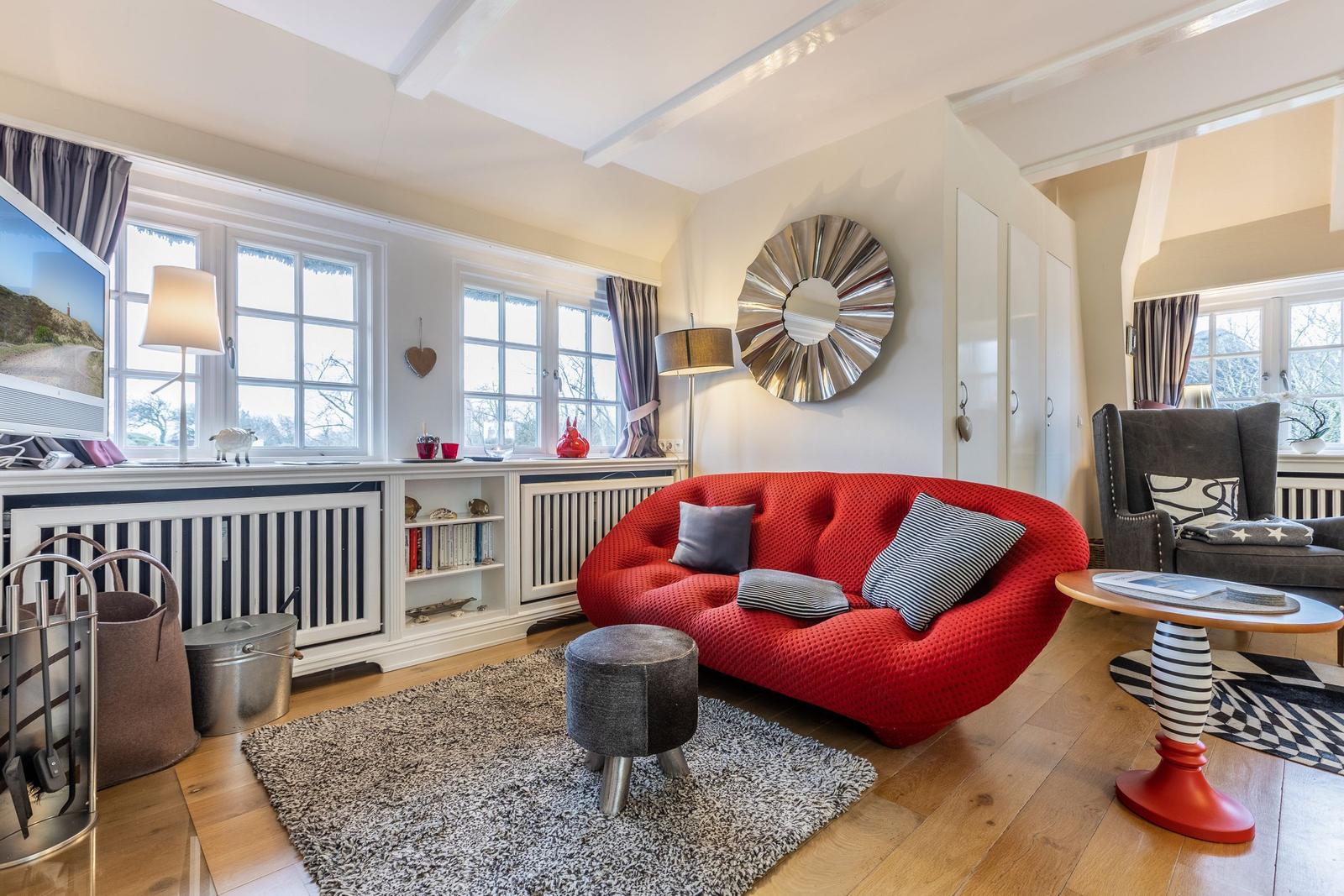 Ein Wohnzimmer mit rotem Sofa, Holzboden und Fenstern mit Vorhängen.