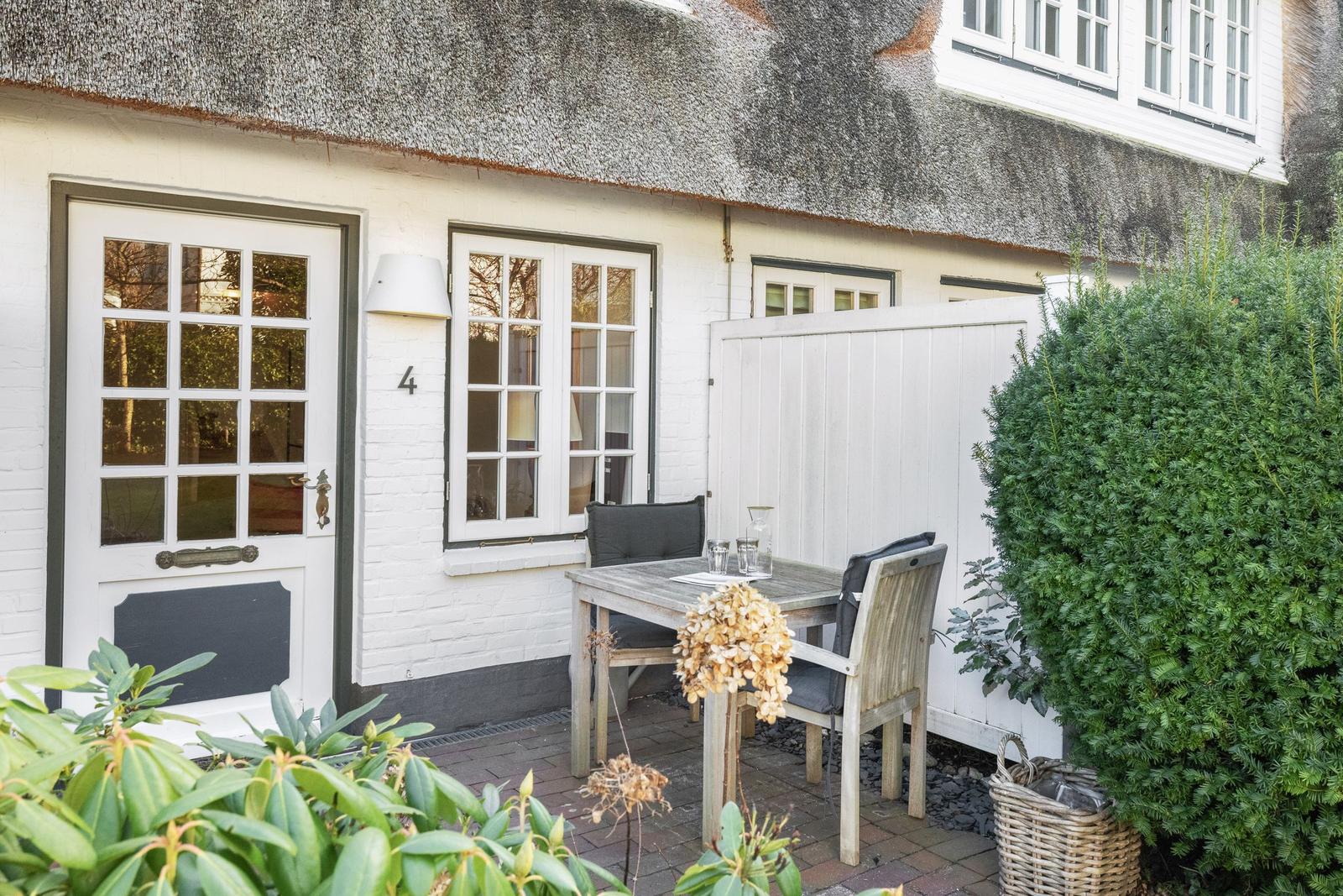 House with thatched roof, white door and windows, wooden table with chairs in the garden.