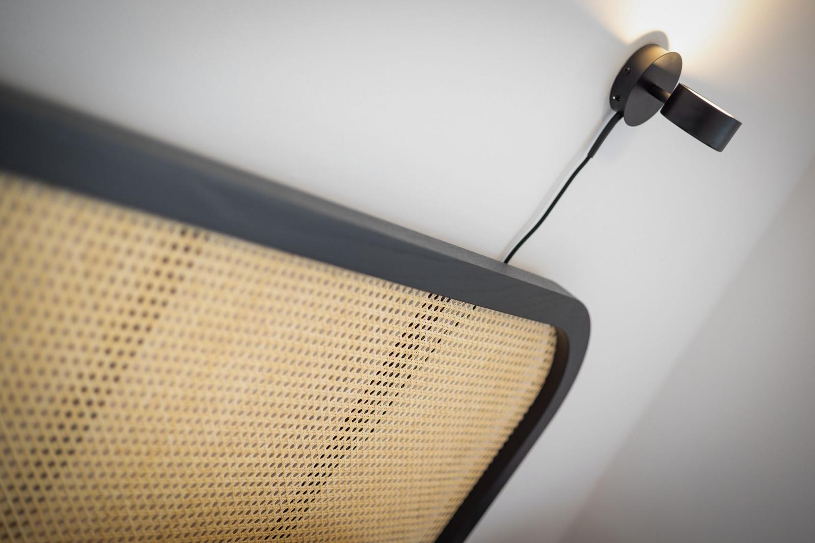 Black wall lamp next to a bed with woven headboard.
