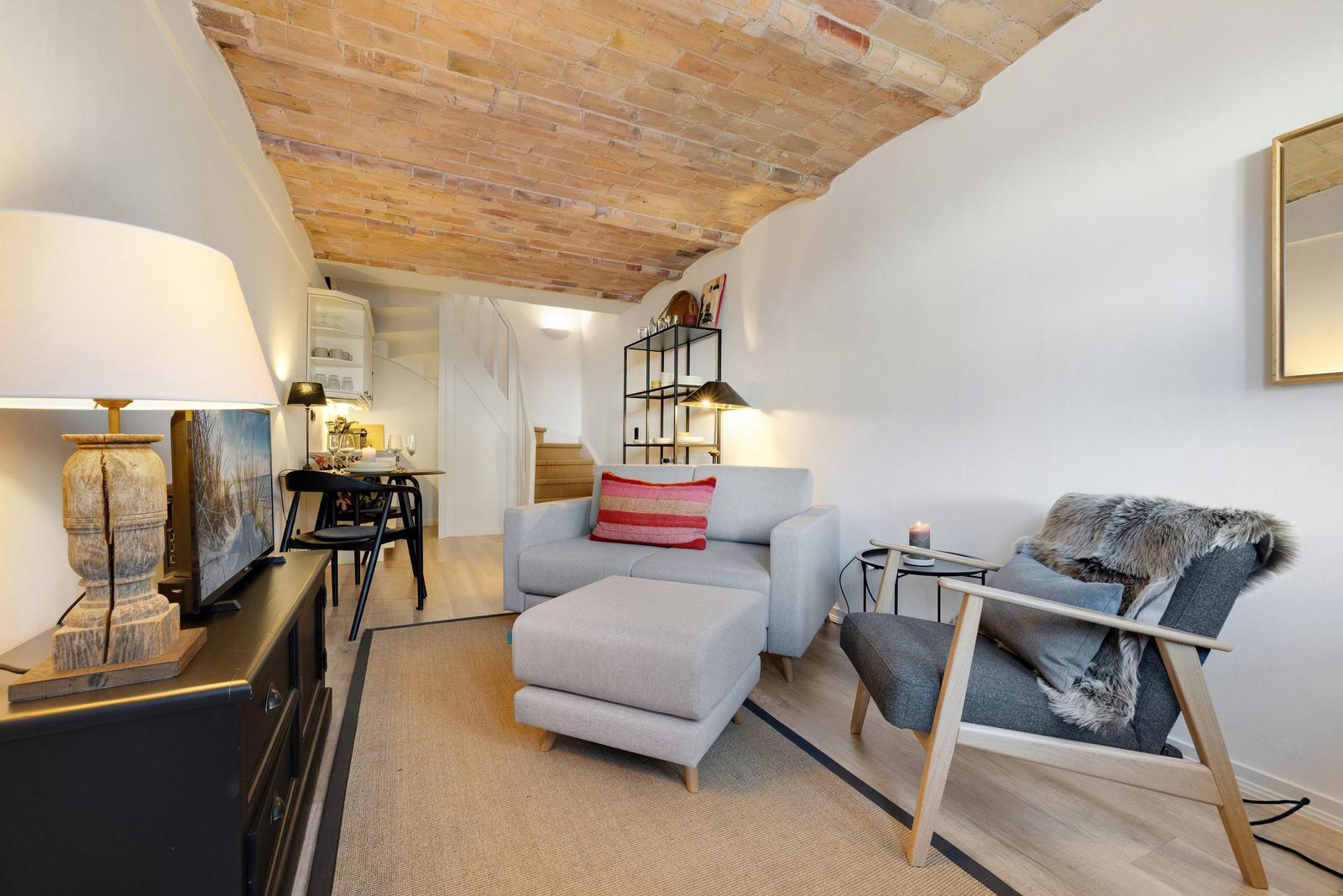 Living room with sofa, armchair, and brick ceiling. Kitchen in the background.