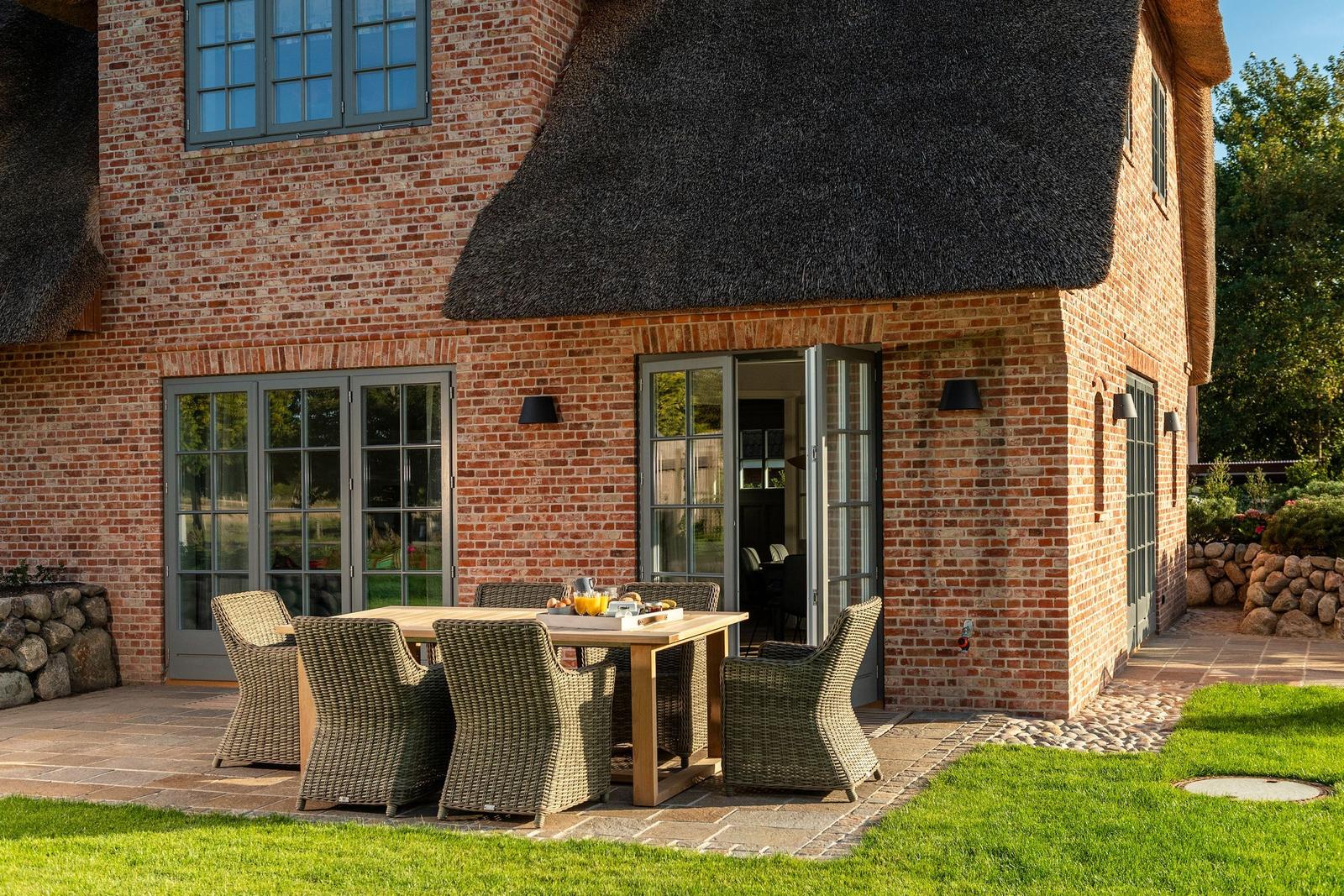 Terrasse mit Esstisch und Stühlen vor einem Backsteinhaus mit Strohdach.