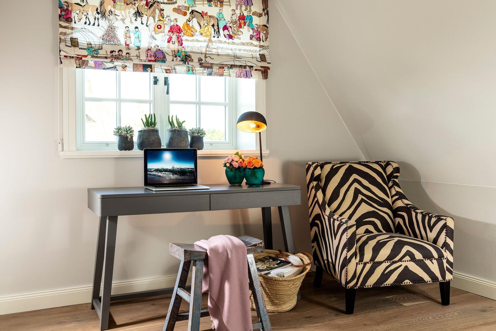 Schreibtisch mit Laptop, Stuhl und Zebra-Stuhl unter Fenster mit Pflanzen.