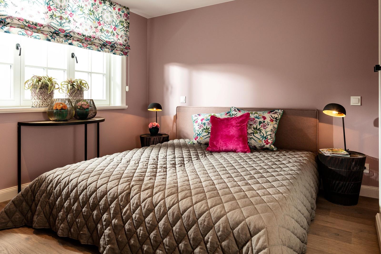 Schlafzimmer mit Bett, Kissen, Nachttischen und Fenster mit Blumenrollo.