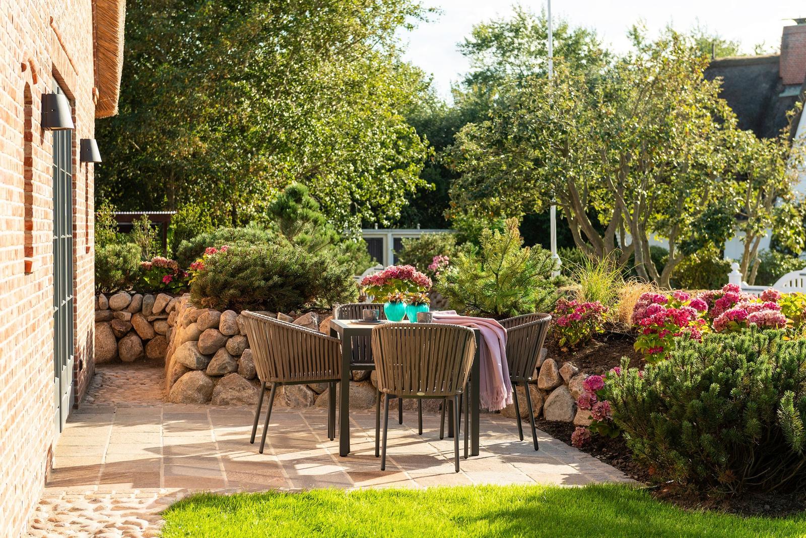 Terrasse mit Tisch, Stühlen und Blumenbeete. Steinmauer und grüne Bäume umgeben den Außenbereich.