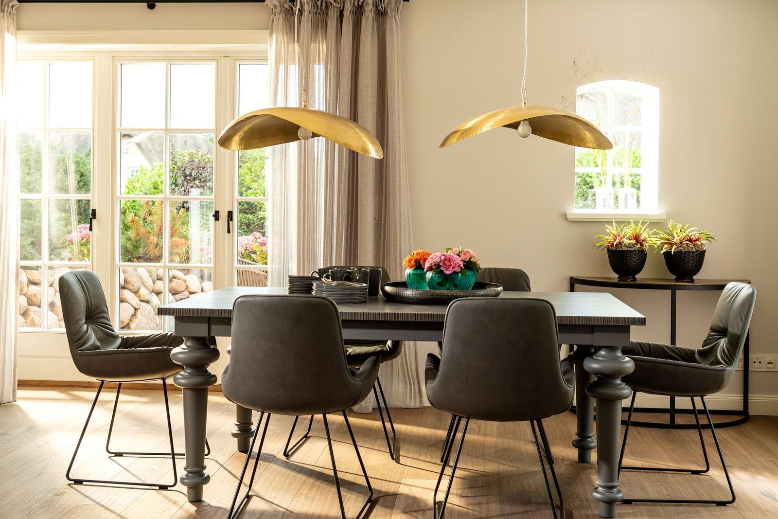 Esszimmer mit Tisch, Stühlen und goldenen Deckenleuchten. Fenster mit Blick auf Garten.