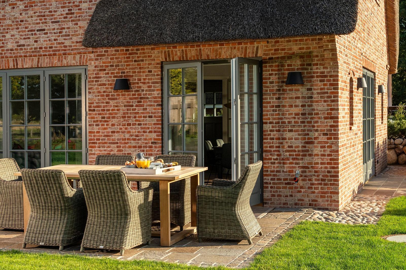 Terrasse mit Holztisch und Stühlen vor einem Backsteinhaus mit Dach aus Stroh.