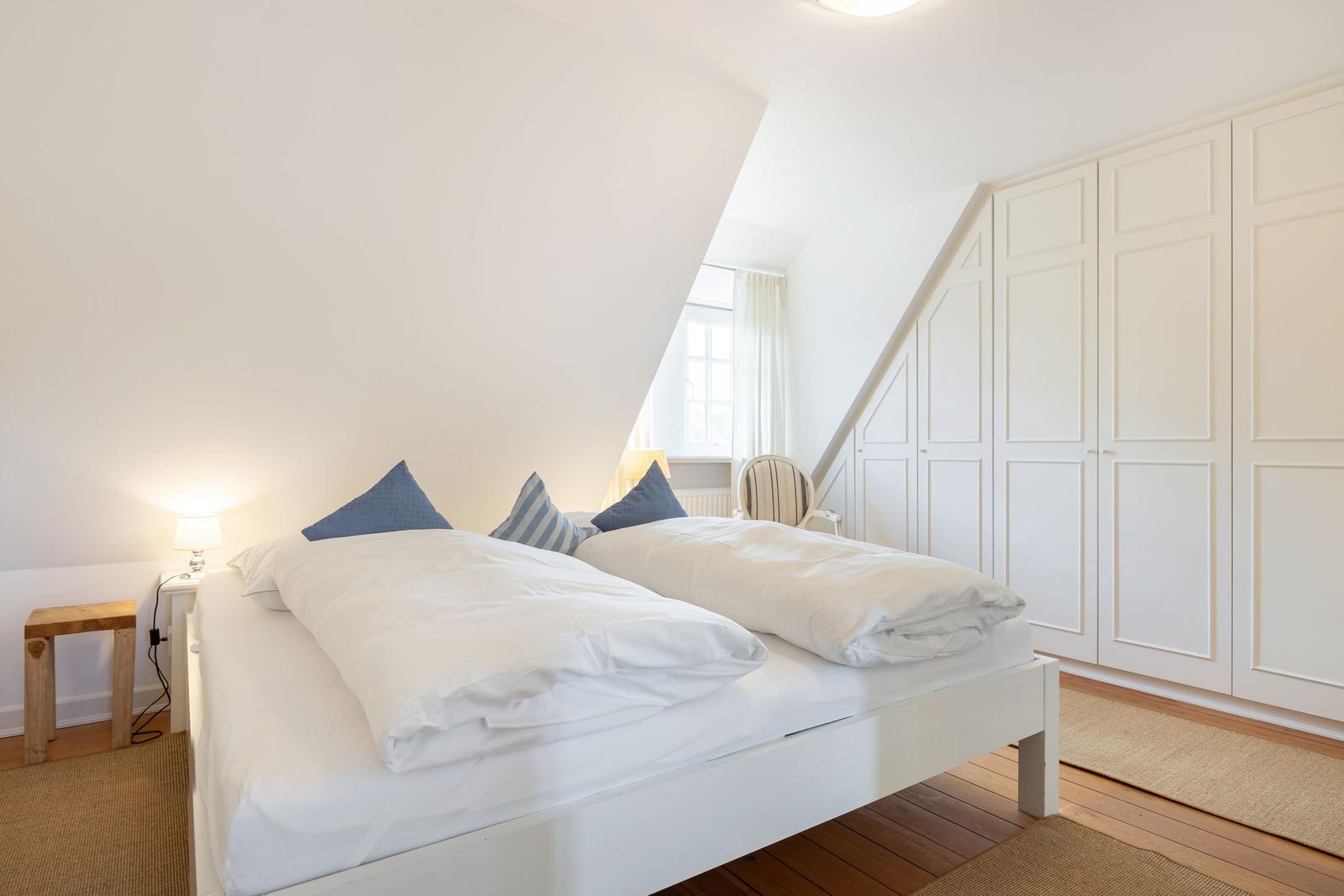 Schlafzimmer mit Doppelbett, weißen Wänden und Holzboden. An der Wand steht ein weißer Schrank.