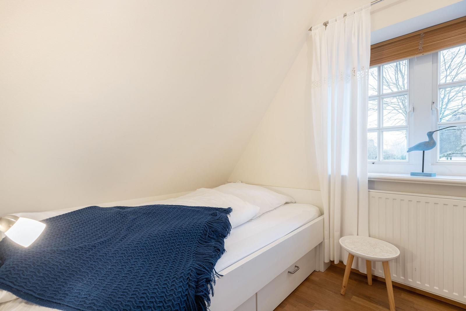 Schlafzimmer mit Dachschräge, Bett mit blauer Decke und Fenster mit Vorhängen.
