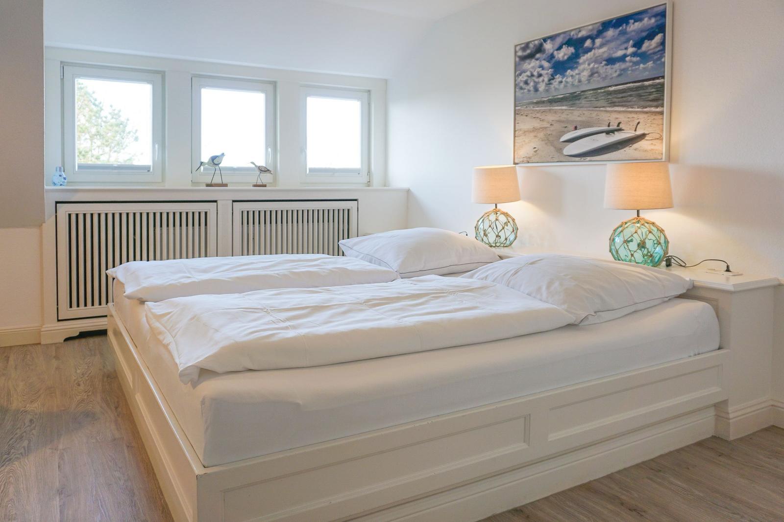 Schlafzimmer mit weißem Bett, zwei Nachttischen und Fenstern mit Blick nach draußen.
