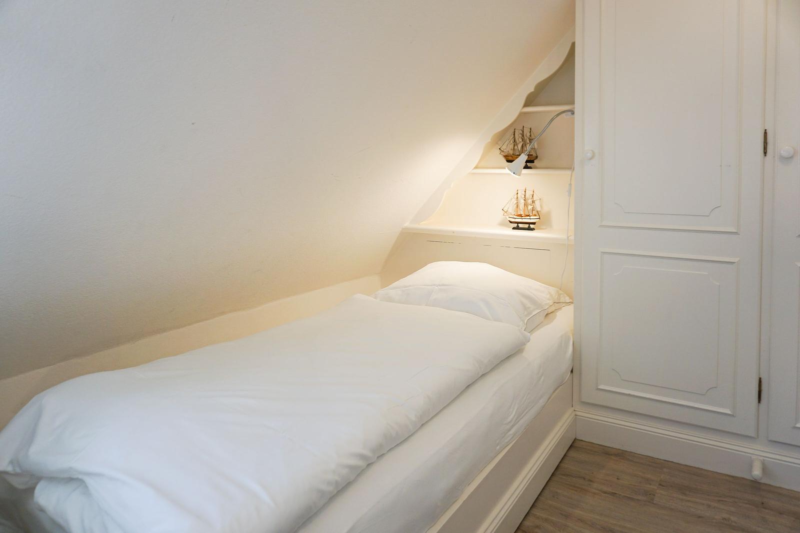 Schlafzimmer mit Einzelbett, weißen Wänden und schräger Decke. Weiße Schrankwand und Nachttisch mit Lampe.
