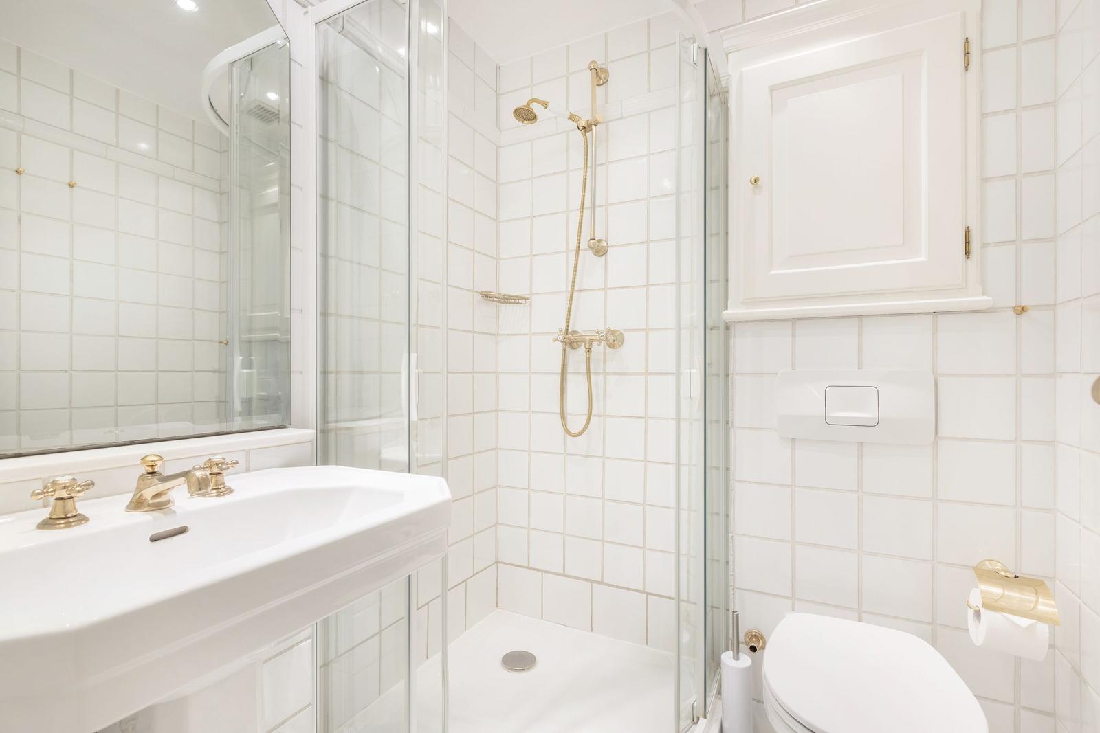 Badezimmer mit Dusche, Waschbecken und Toilette. Weiße Fliesen und goldene Armaturen.