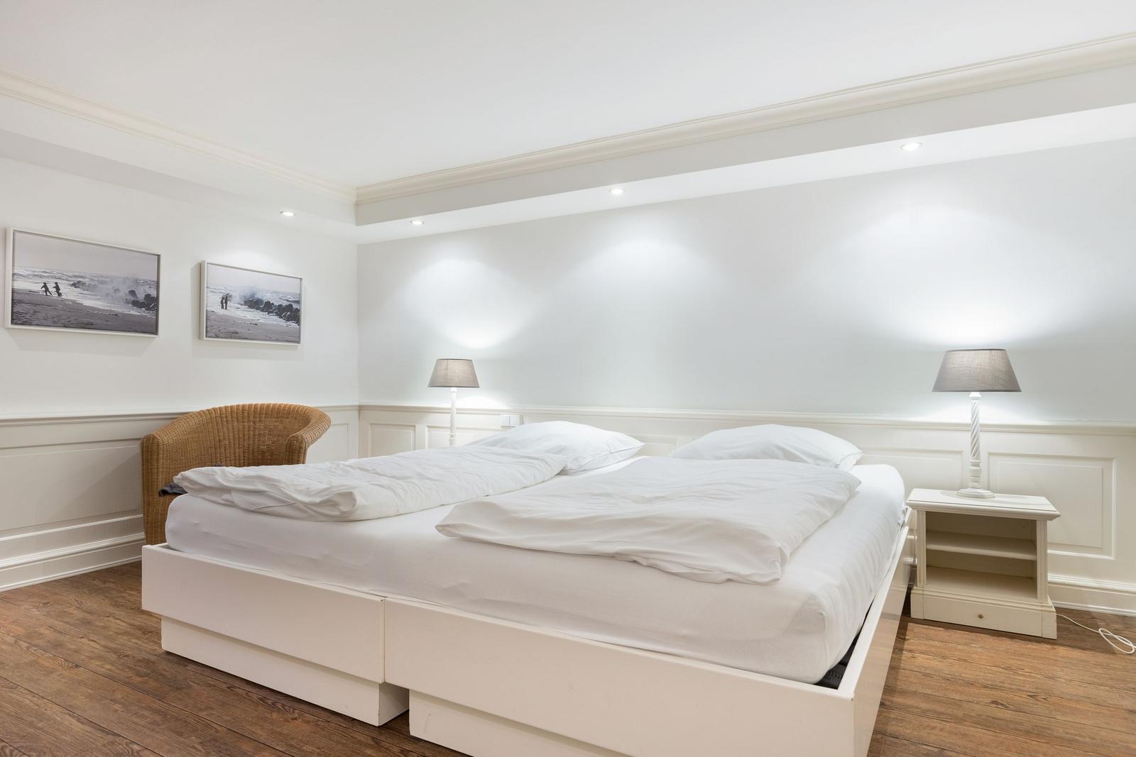 Schlafzimmer mit weißem Bett, Nachttisch und Lampe. Wandbilder und Holzboden.