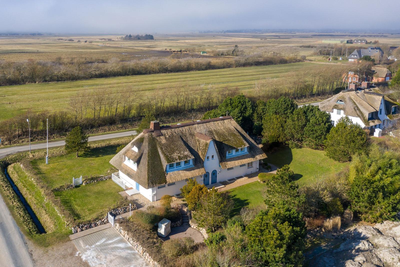 Dorfhaus mit Strohdach und Garten, umgeben von Feldern und Bäumen.