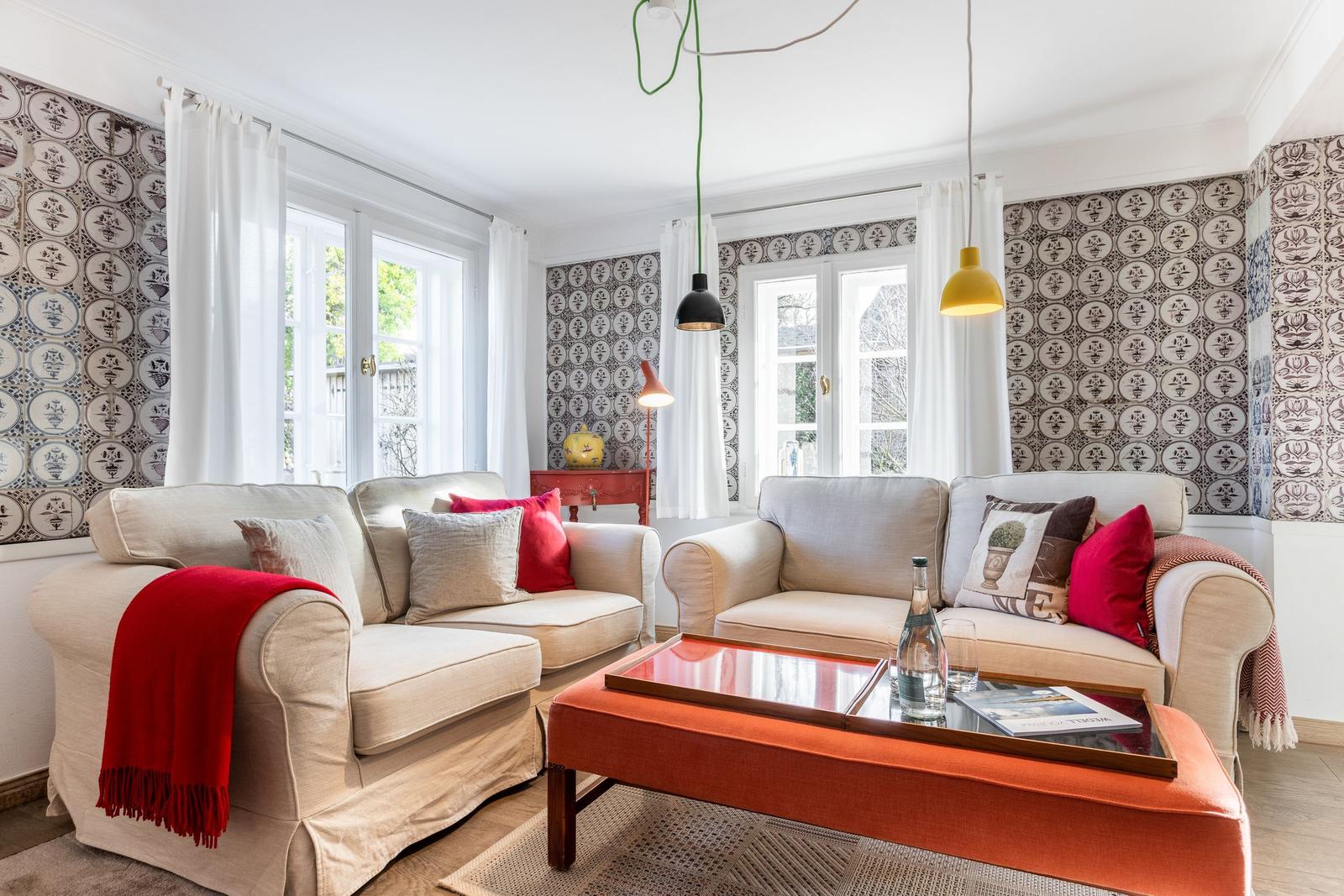 Two sofas with cushions and blanket, table with glass top, windows with white curtains and patterned walls.
