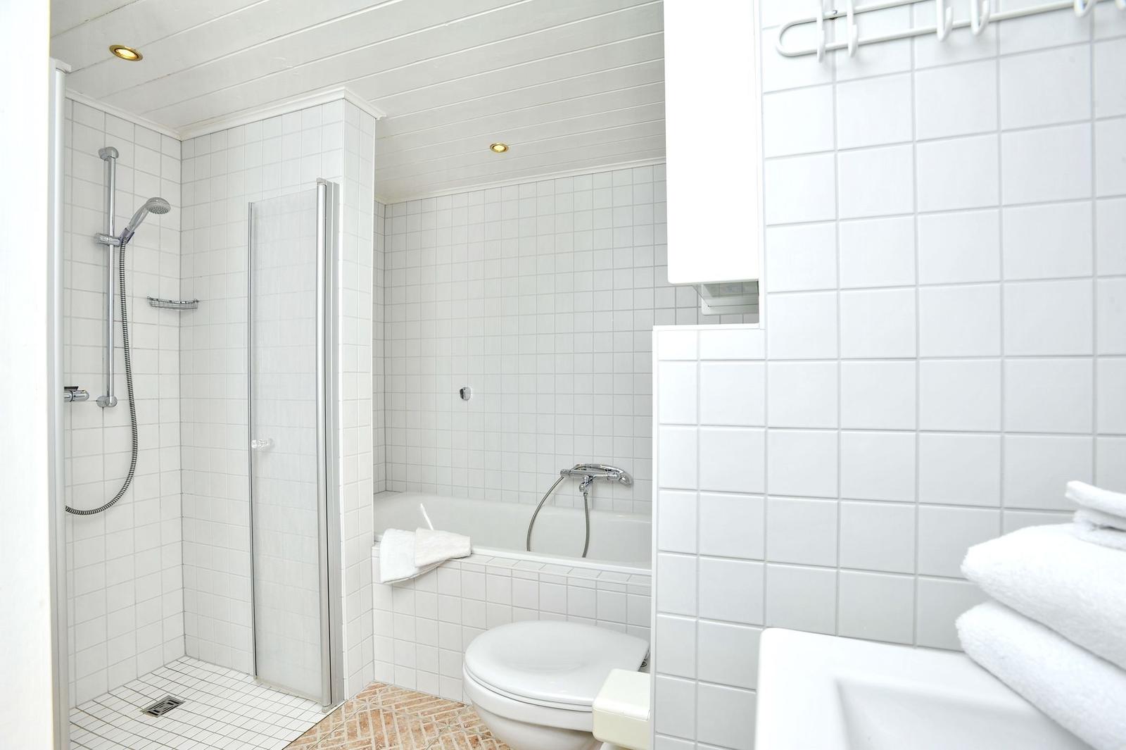 Badezimmer mit Dusche, Badewanne und Toilette. Weiße Fliesen und Handtücher.