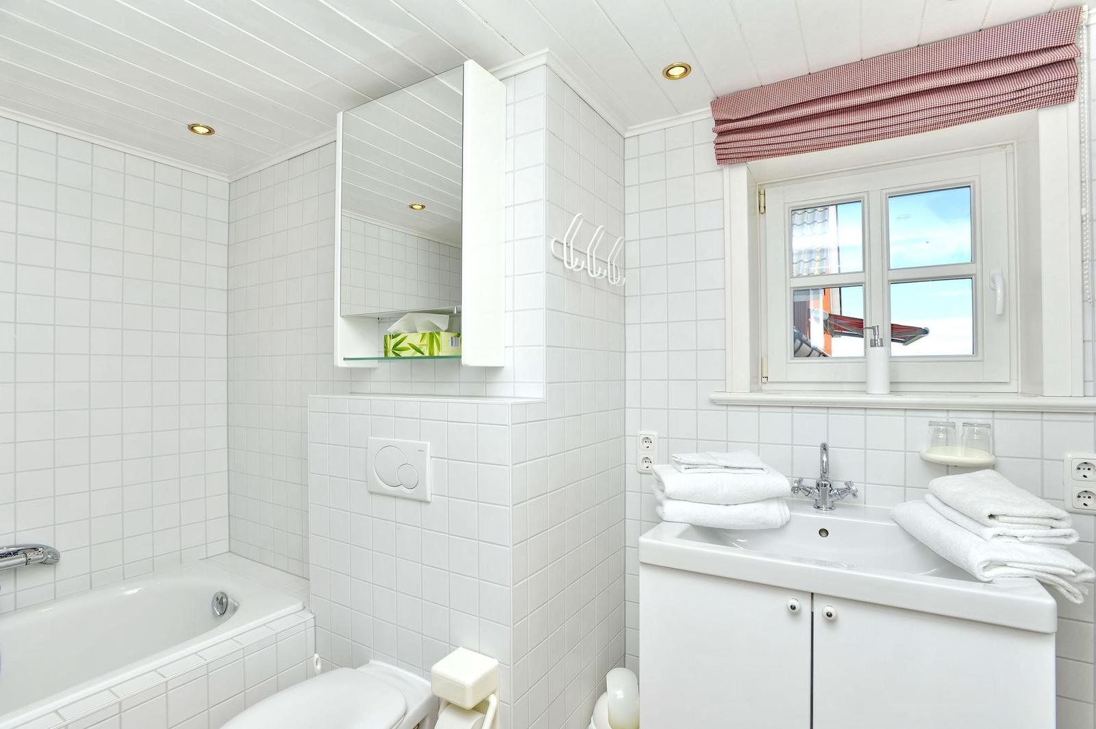 Badezimmer mit Badewanne, Waschbecken und Toilette. Weiße Fliesen und Fenster mit Rollladen.