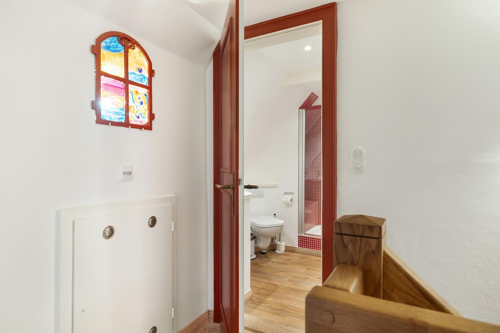 Hallway with colorful window, door to bathroom with toilet and shower.