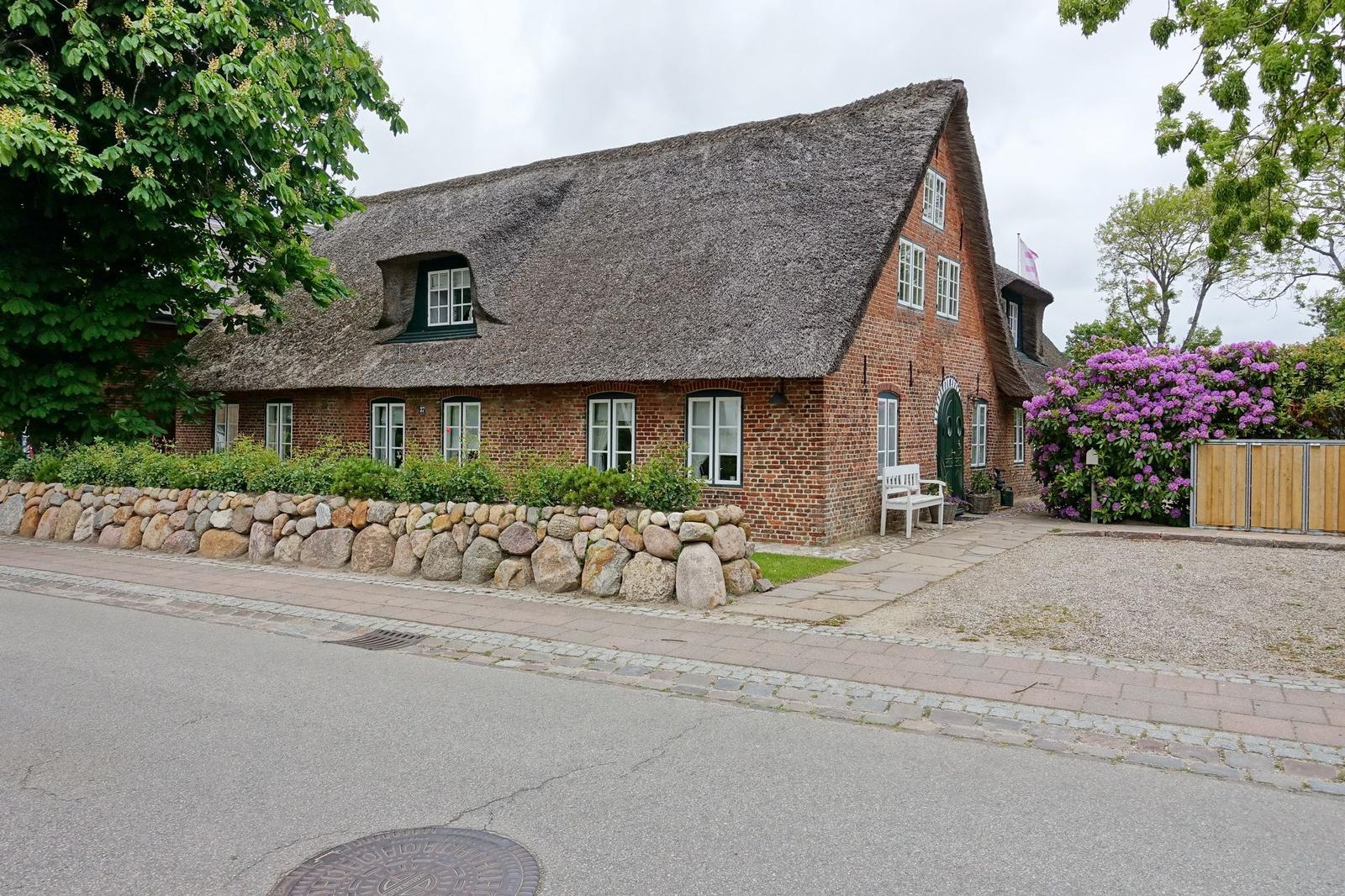 Red brick house with thatched roof, white windows, stone wall, and flowering shrubs.