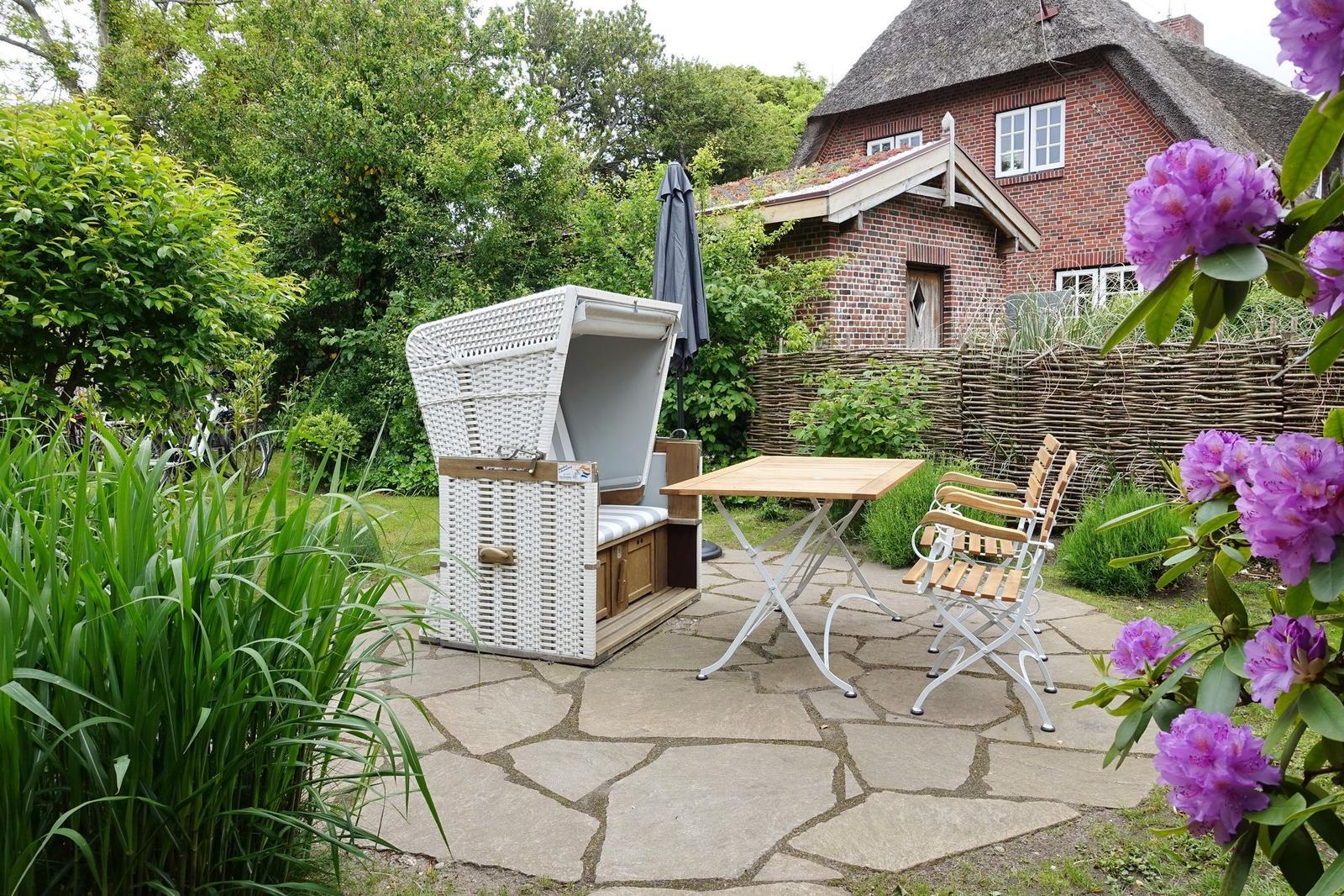 Terrace with white lounge chair, table and chairs. Background: house with reed fence and flowers.