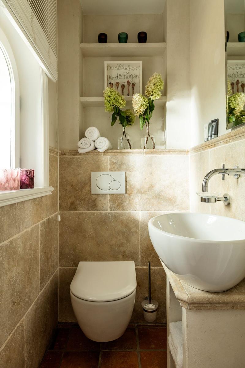 Badezimmer mit Toilette, Waschbecken und Fenster. Wandregale mit Blumen und Handtüchern.