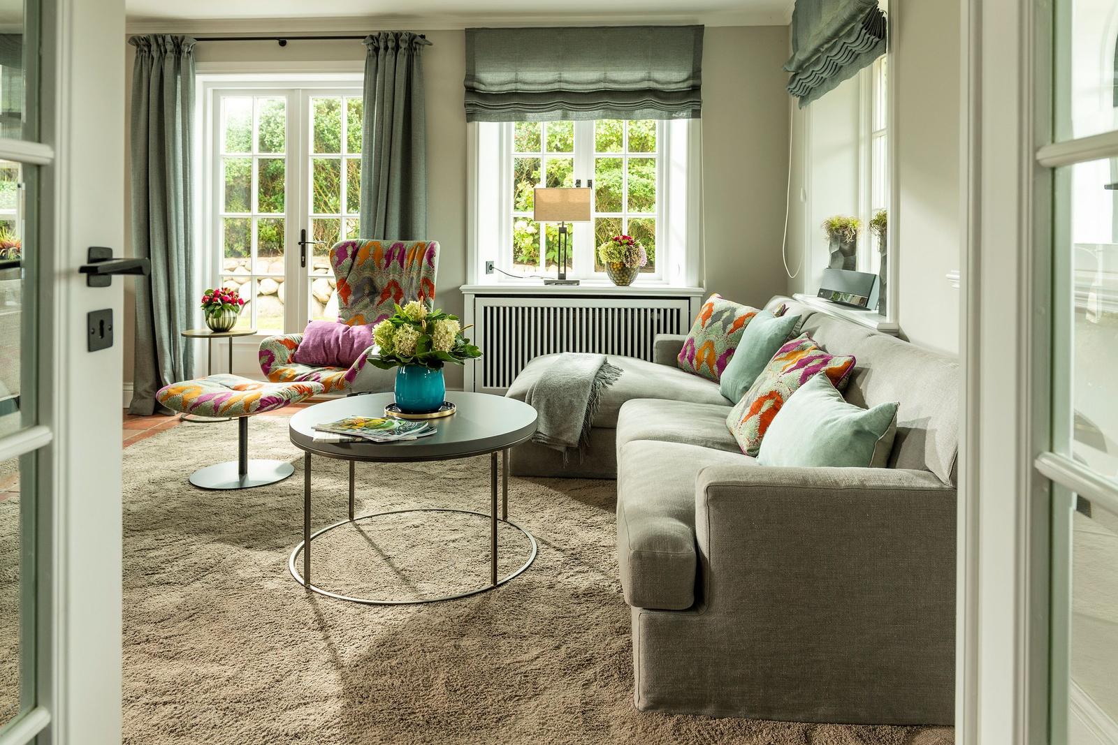 Ein Wohnzimmer mit Sofa, Sessel und Tisch. Fenster mit Vorhängen und Blumen.