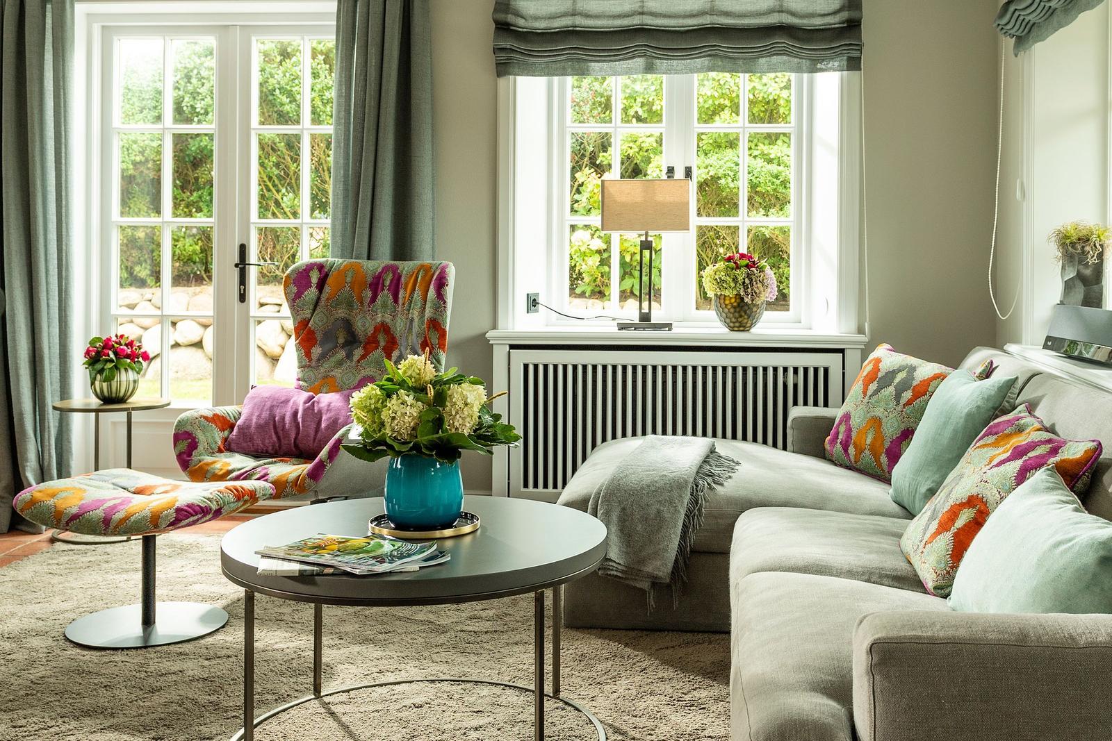 Ein Wohnzimmer mit Sofa, Sessel und Tisch. Fenster und Vorhänge sind sichtbar.