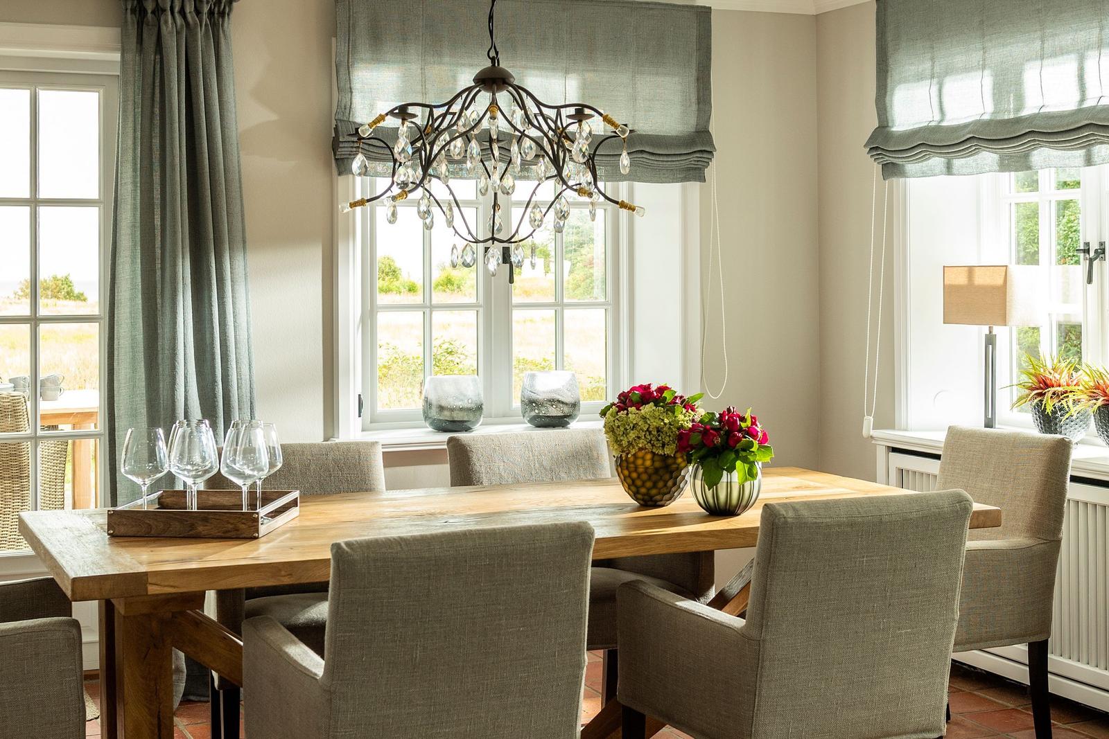 Esszimmer mit Holztisch, Stühlen und Kronleuchter. Fenster mit Rollläden und Blumenarrangements.