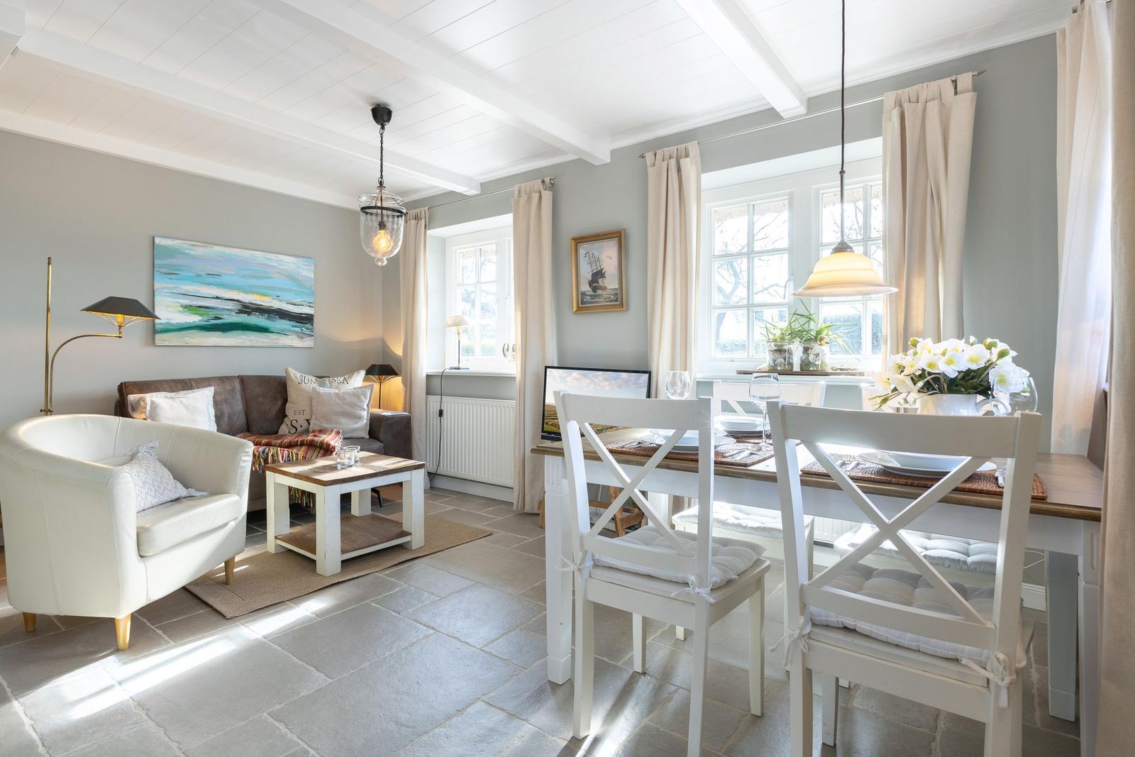 Living and dining area with white table, chairs, and sofa. Stone floor and large windows.