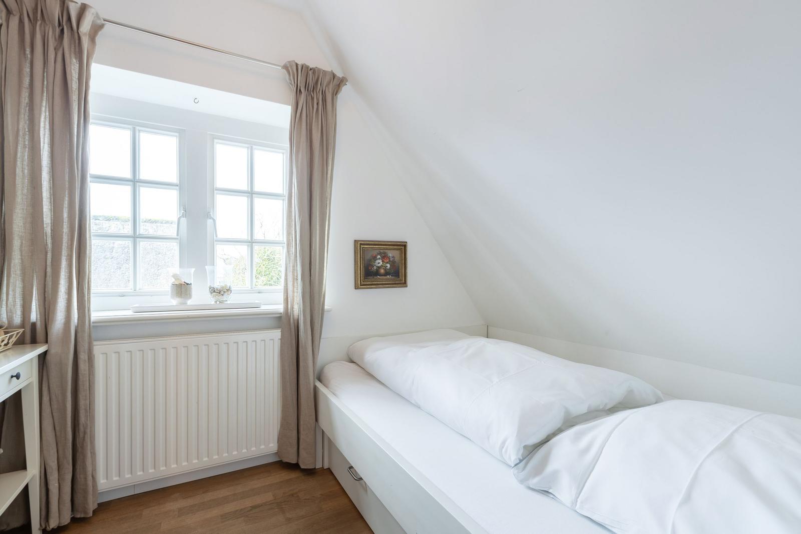 A bedroom with sloped ceiling, white bed, and window with curtains.