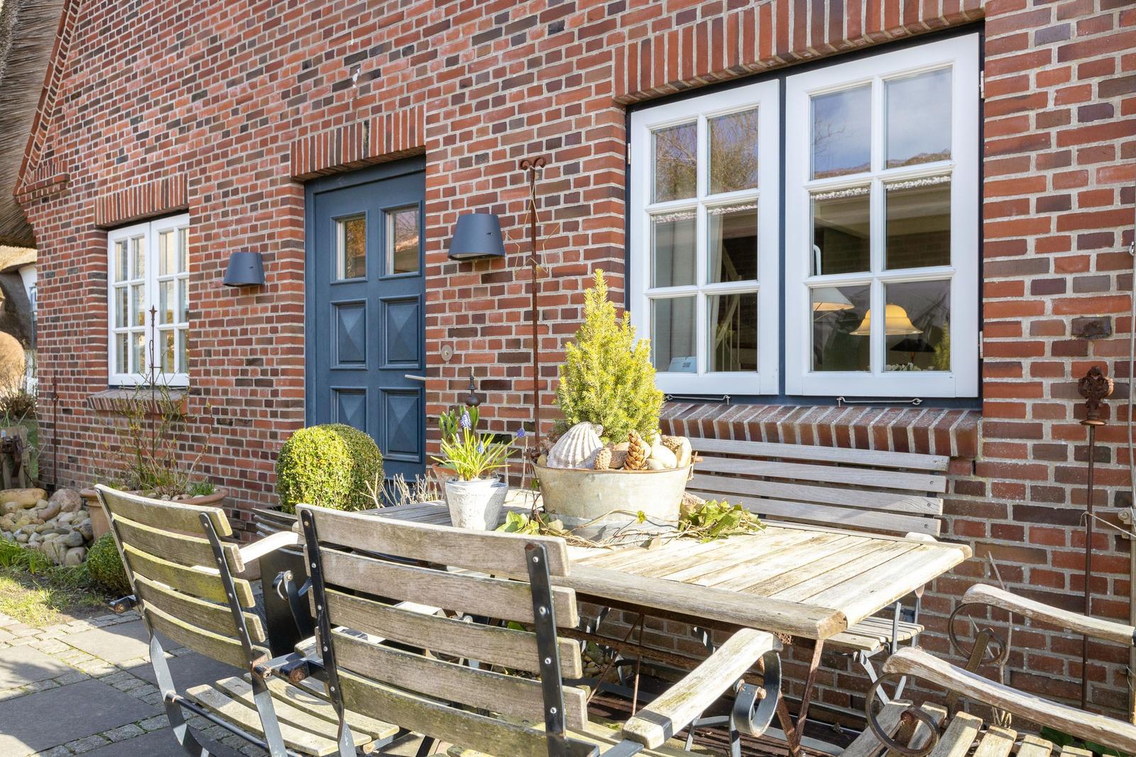 Wooden table and chairs in front of red brick house with blue door and white windows.