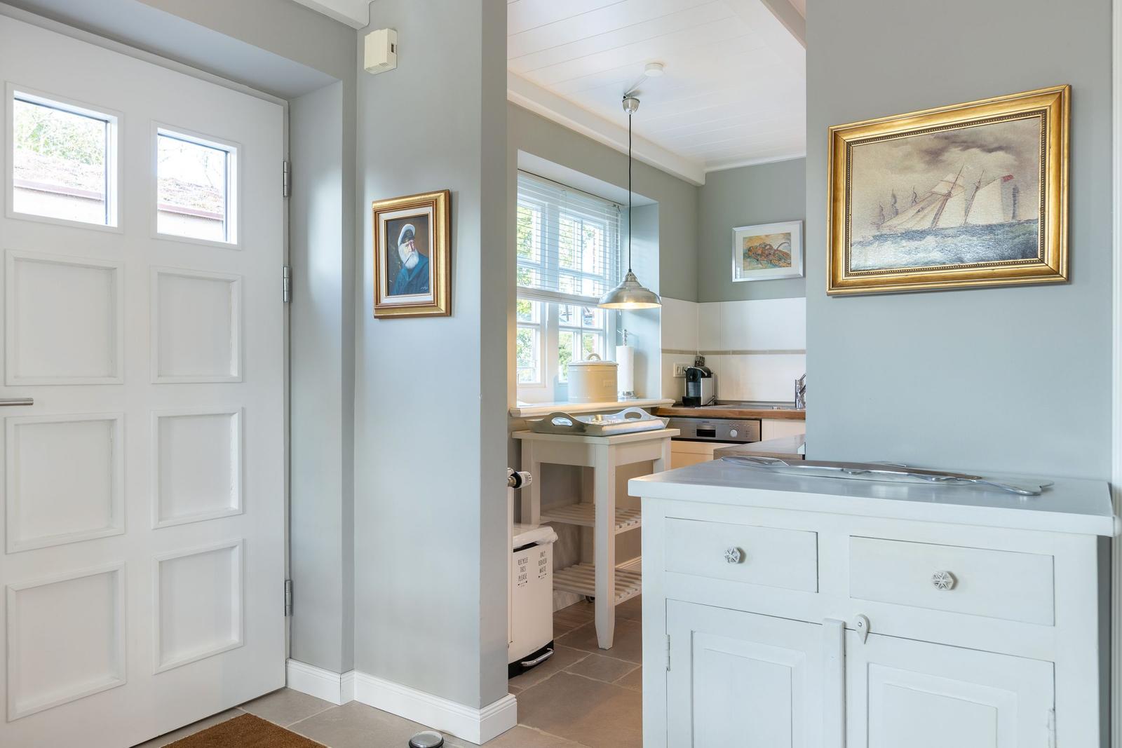 House with kitchen area and entrance door. Wall pictures and white furniture.