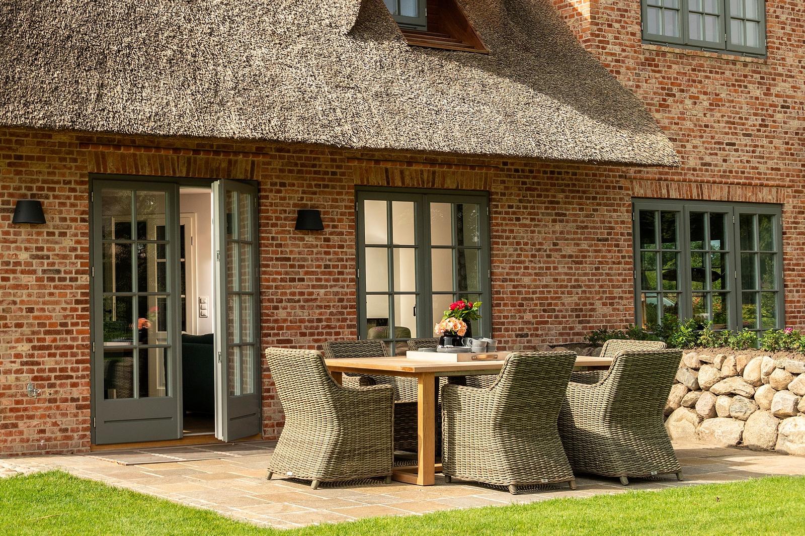Haus mit Strohdach, Terrasse mit Tisch und Stühlen, Holztisch, Wickerstühle, Steinmauer, Fenster und Tür.