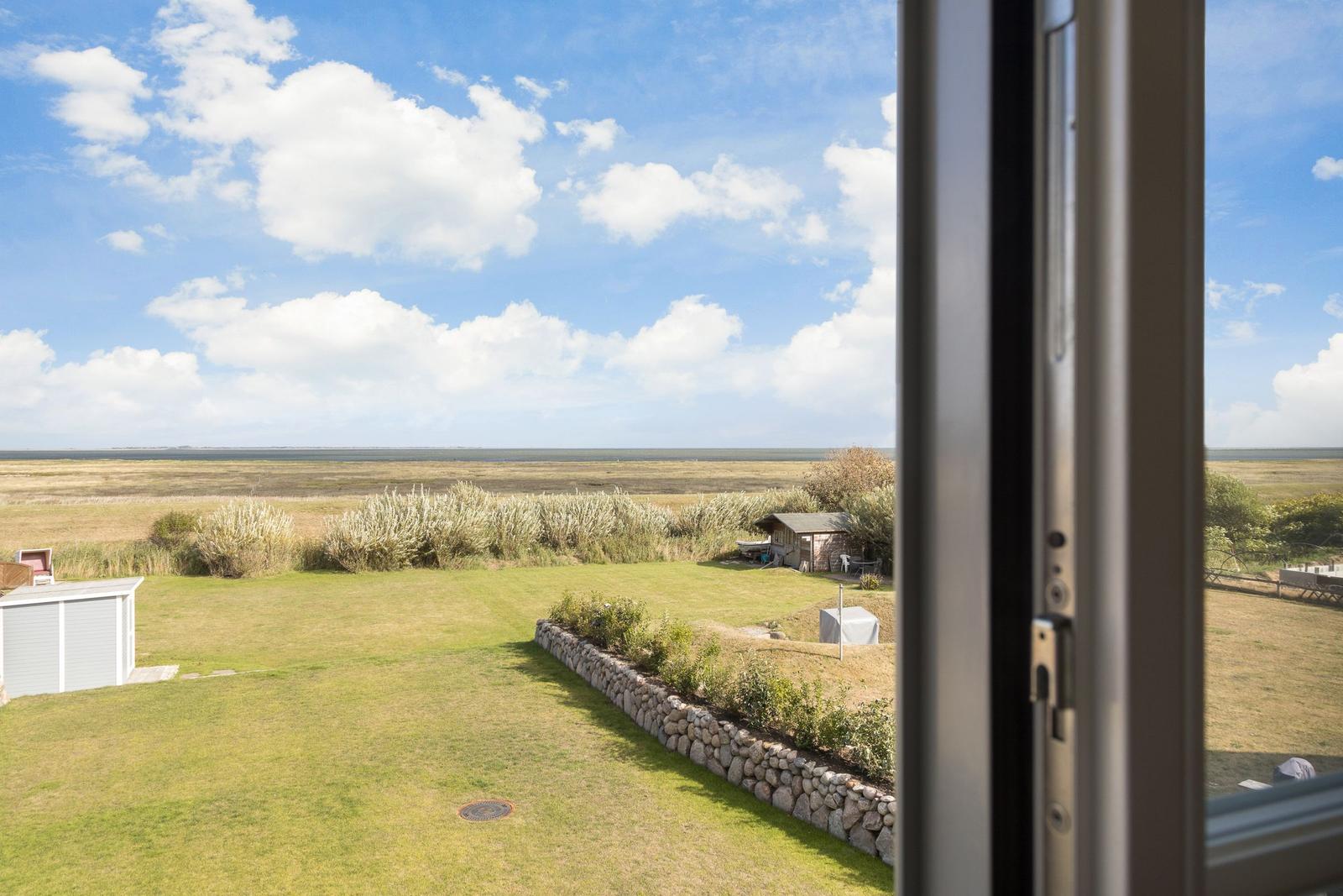 Blick aus dem Fenster auf Grasfläche, Steinmauer und Felder unter blauem Himmel mit weißen Wolken.