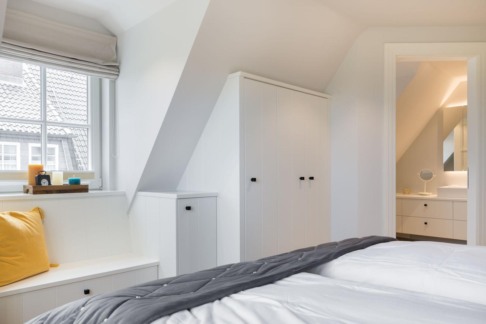 Schlafzimmer mit Dachschrägen, weißem Schrank und Fenster mit Blick auf Dächer.