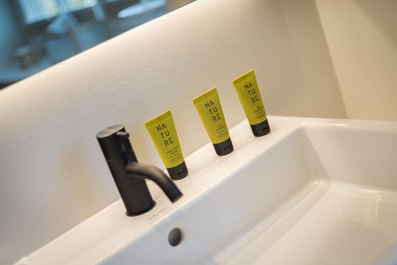 Drei gelbe Natur-Duschgels, Shampoo und Lotion auf einer weißen Waschschale mit schwarzer Wasserhahn.
