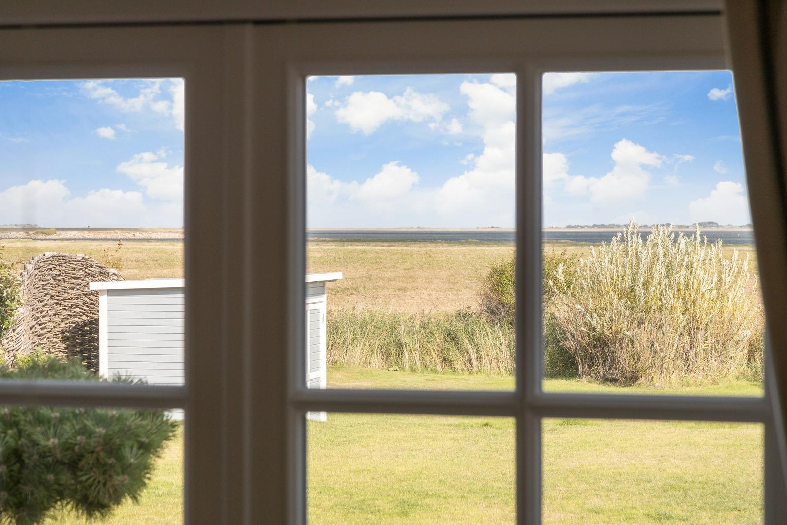 Durch das Fenster blickt man auf eine Wiese mit Gräsern und einen Himmel mit Wolken.