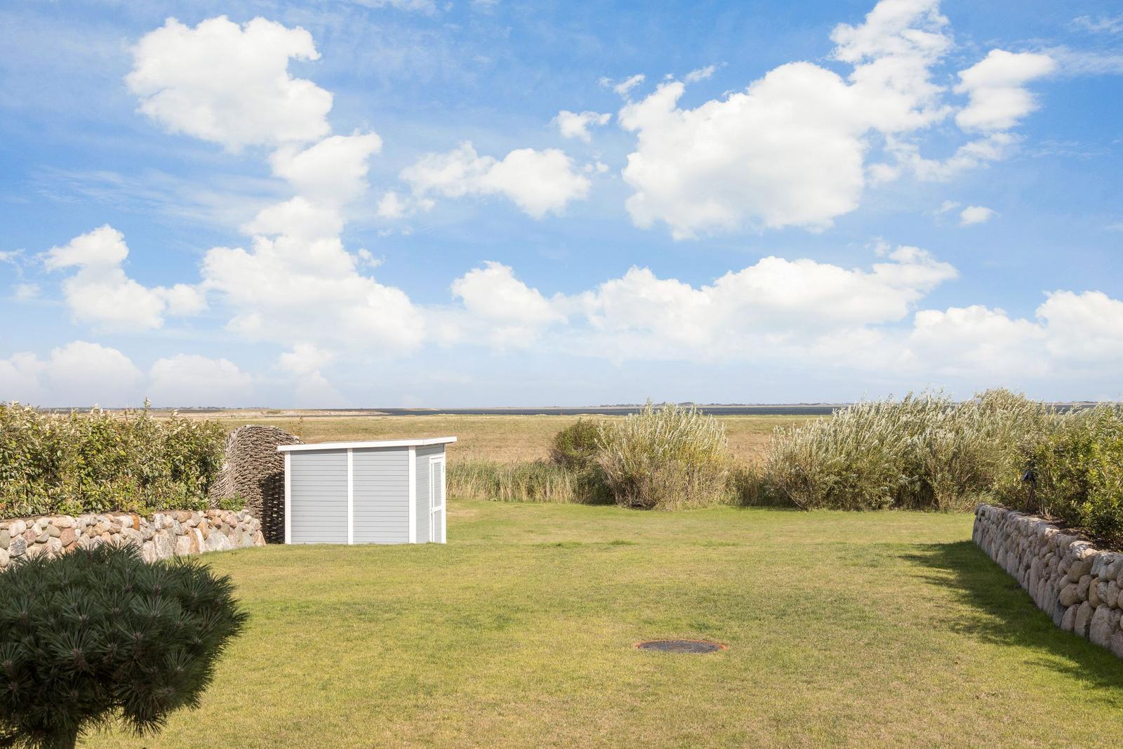 Garten mit Rasenfläche, kleinem Schuppen und Steinmauern unter blauem Himmel mit weißen Wolken.