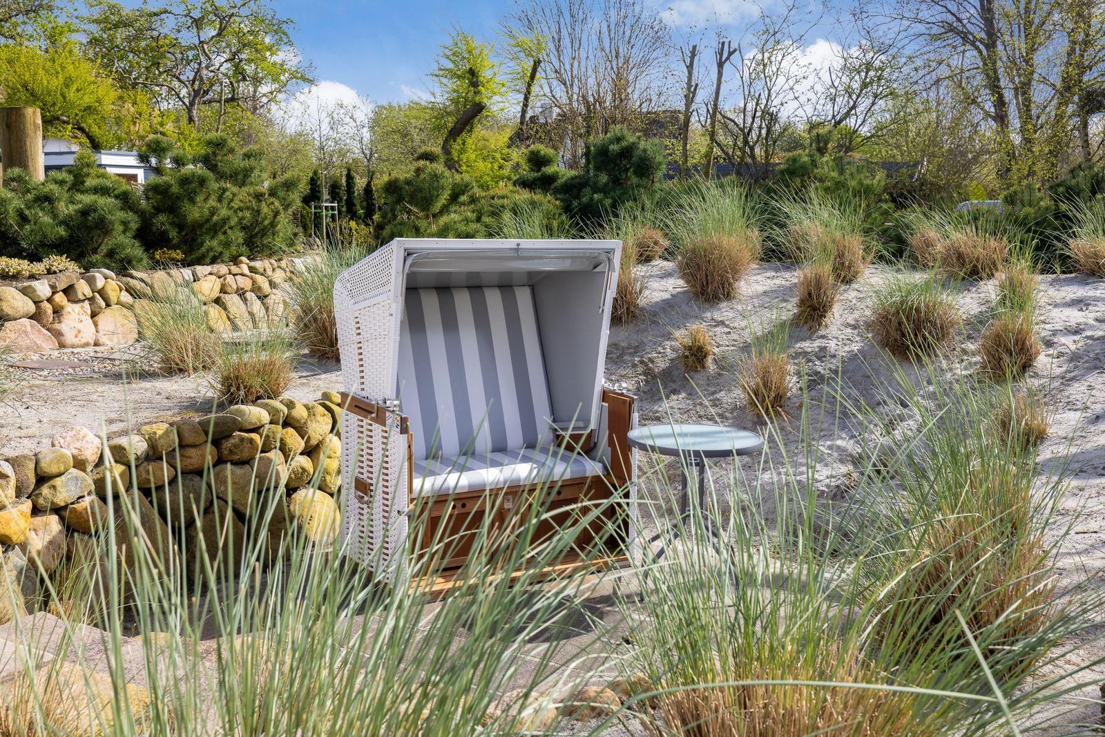 White beach chair with striped cushions on sandy ground with grasses and stone wall.