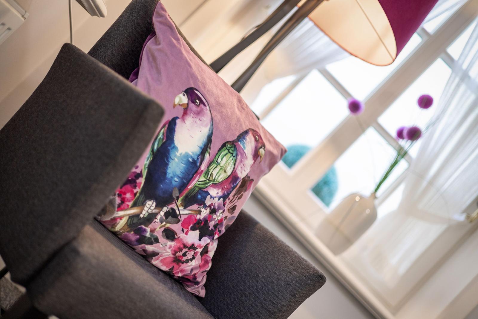 A bird-patterned cushion on a chair near a window with flowers.