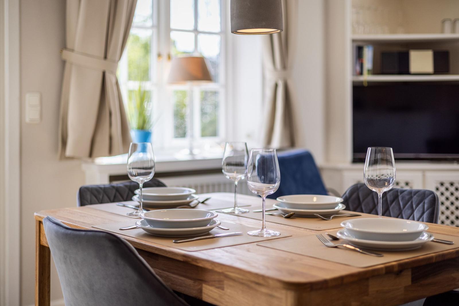 Dining room with wooden table, chairs, and table setting. Window with curtains and TV in background.