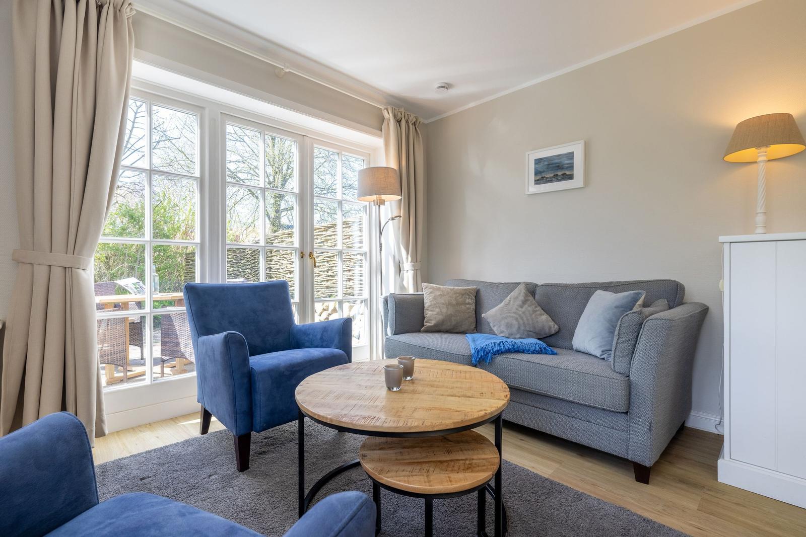 Living room with sofa, blue armchairs, and wooden table. Windows overlook the garden.