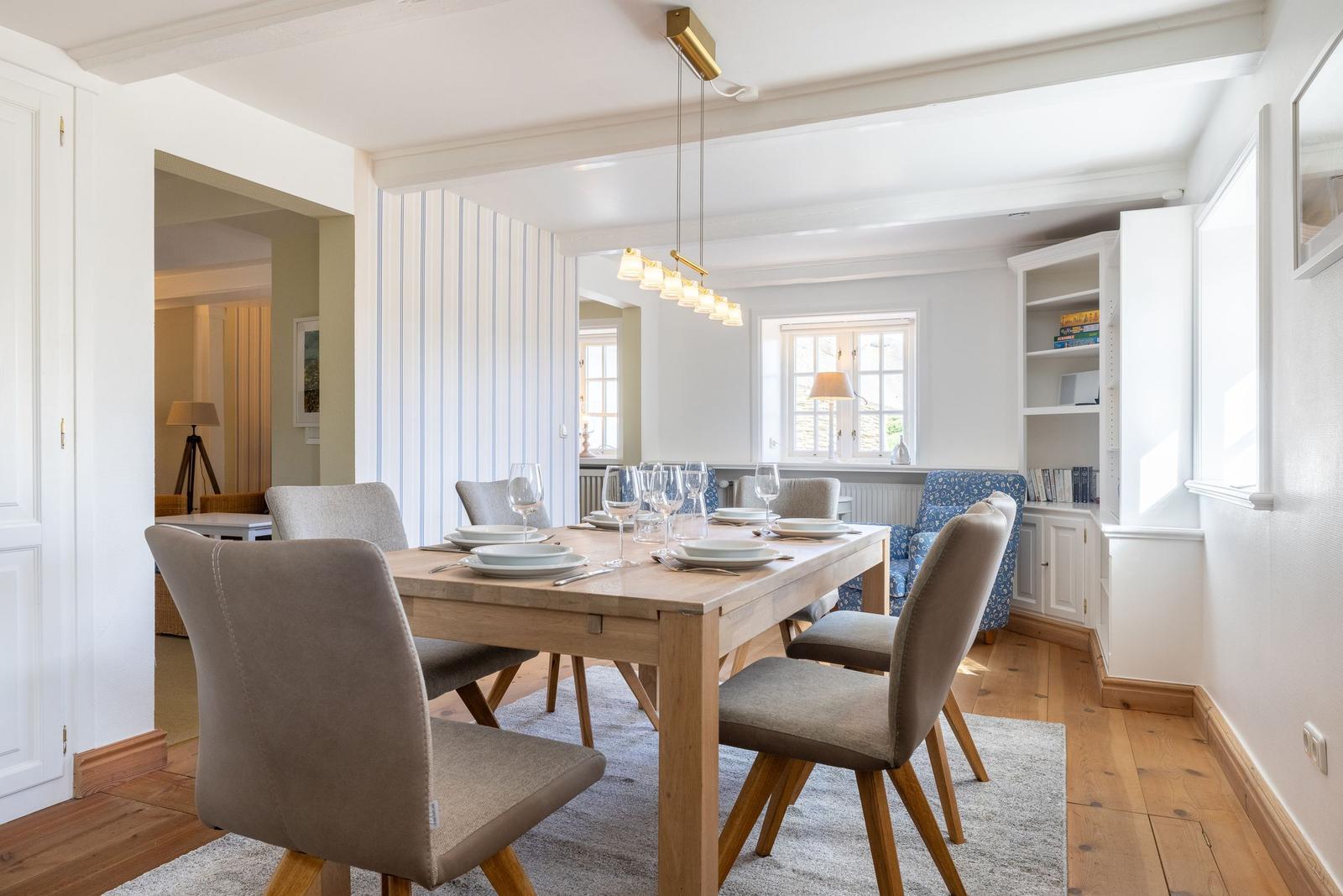 Dining room with wooden table, chairs, and windows. Bookshelf and ceiling light visible.