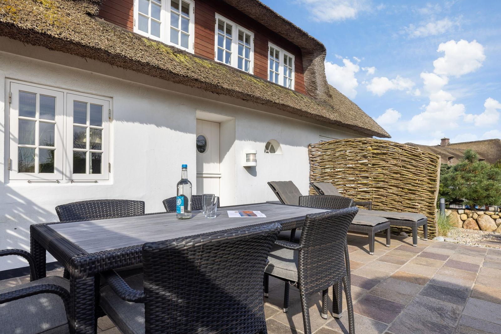 Terrace with table, chairs, and loungers in front of a house with thatched roof.