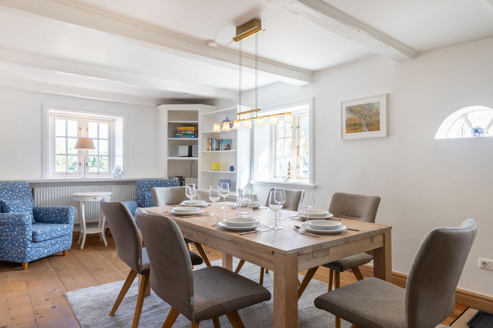 Dining room with wooden table, chairs, and windows. Bookshelf and lamp visible.