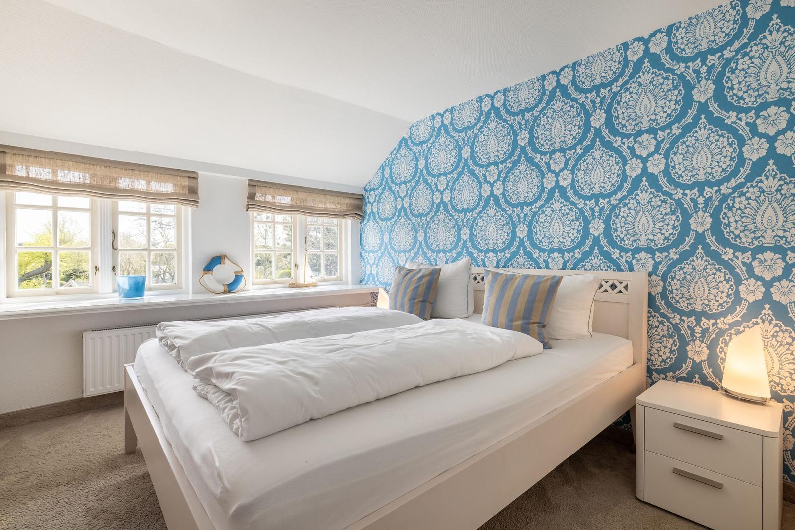 Bedroom with blue-and-white patterned wall, white bed, and windows with outdoor view.