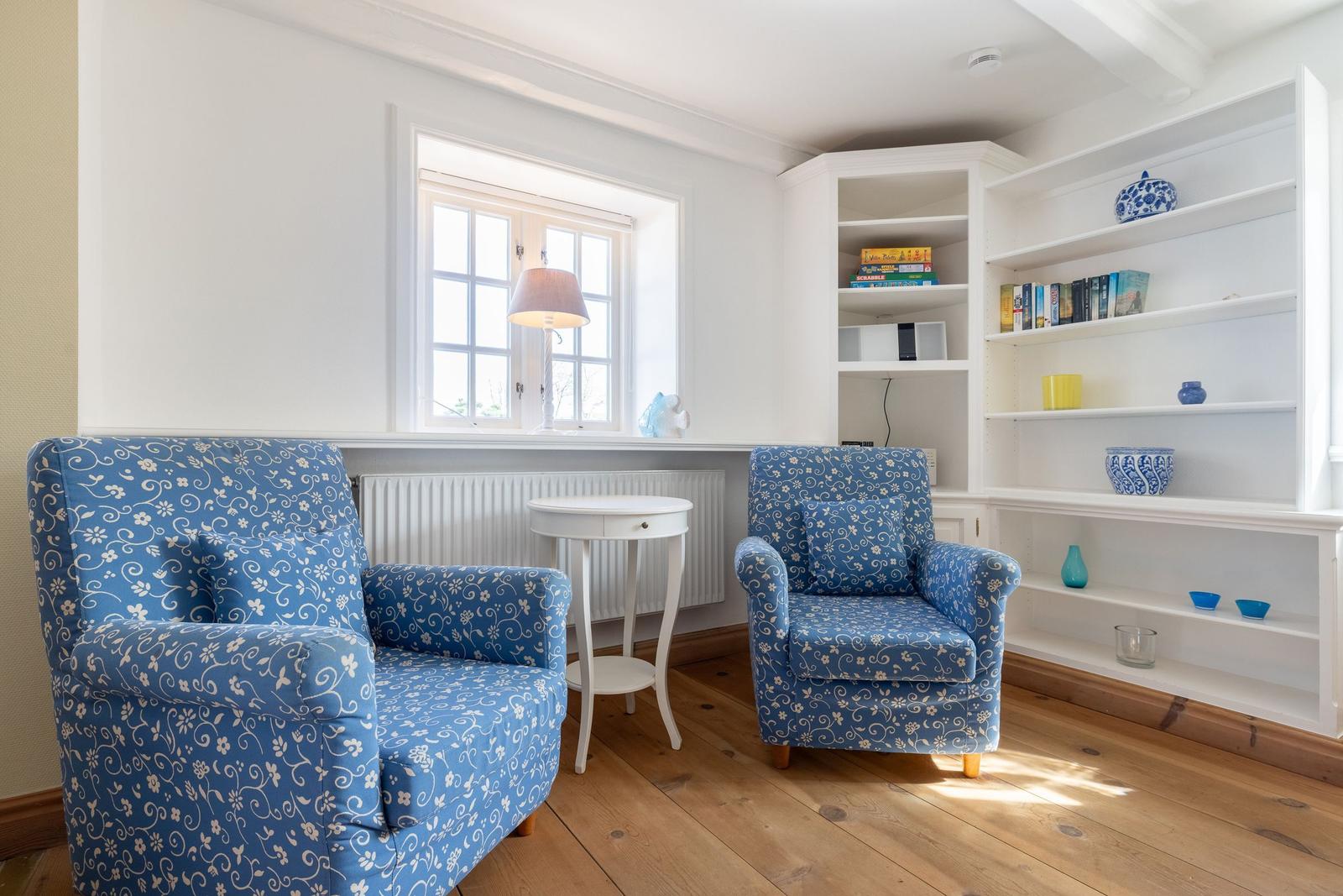 Two blue patterned armchairs are by a window. White shelves are on the wall.