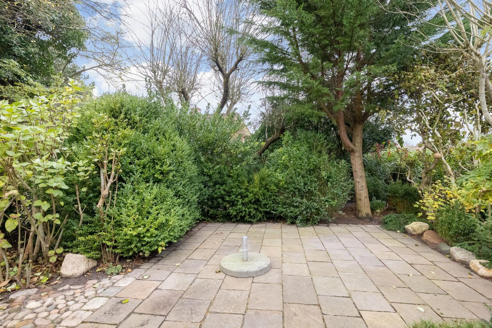 Garden with stone paving, trees, and shrubs. Central metal post on stone base.