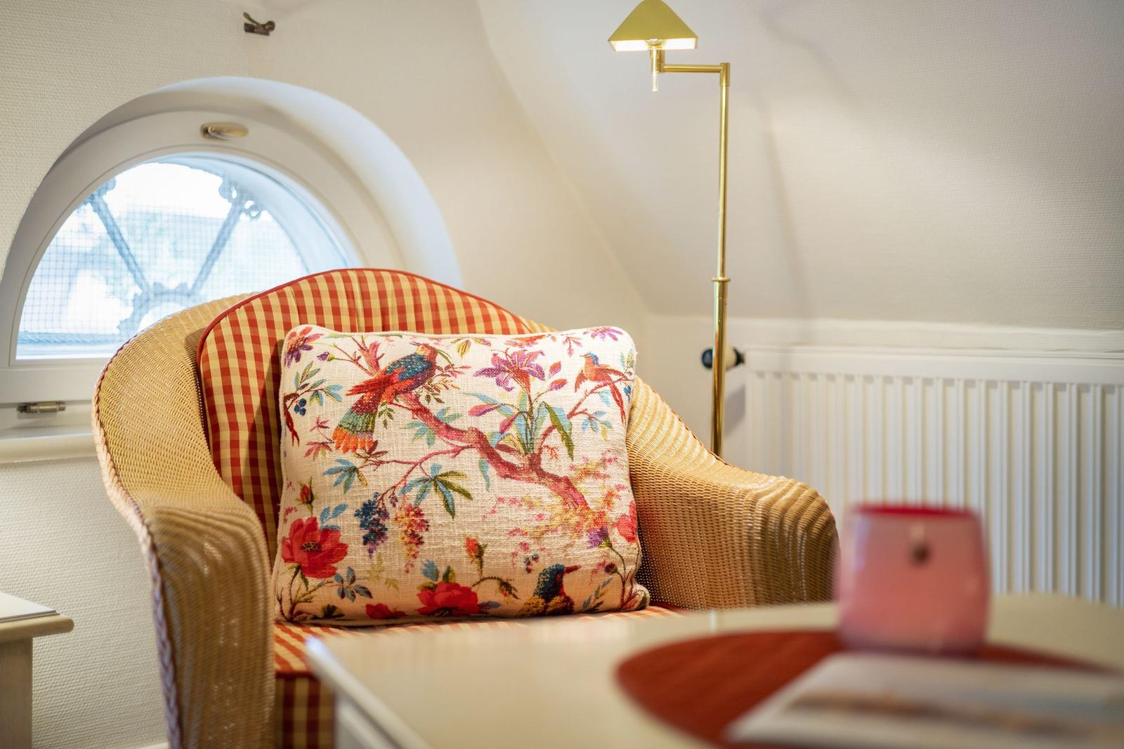 A chair with patterned upholstery and a cushion with bird and flower motifs in front of a round window.