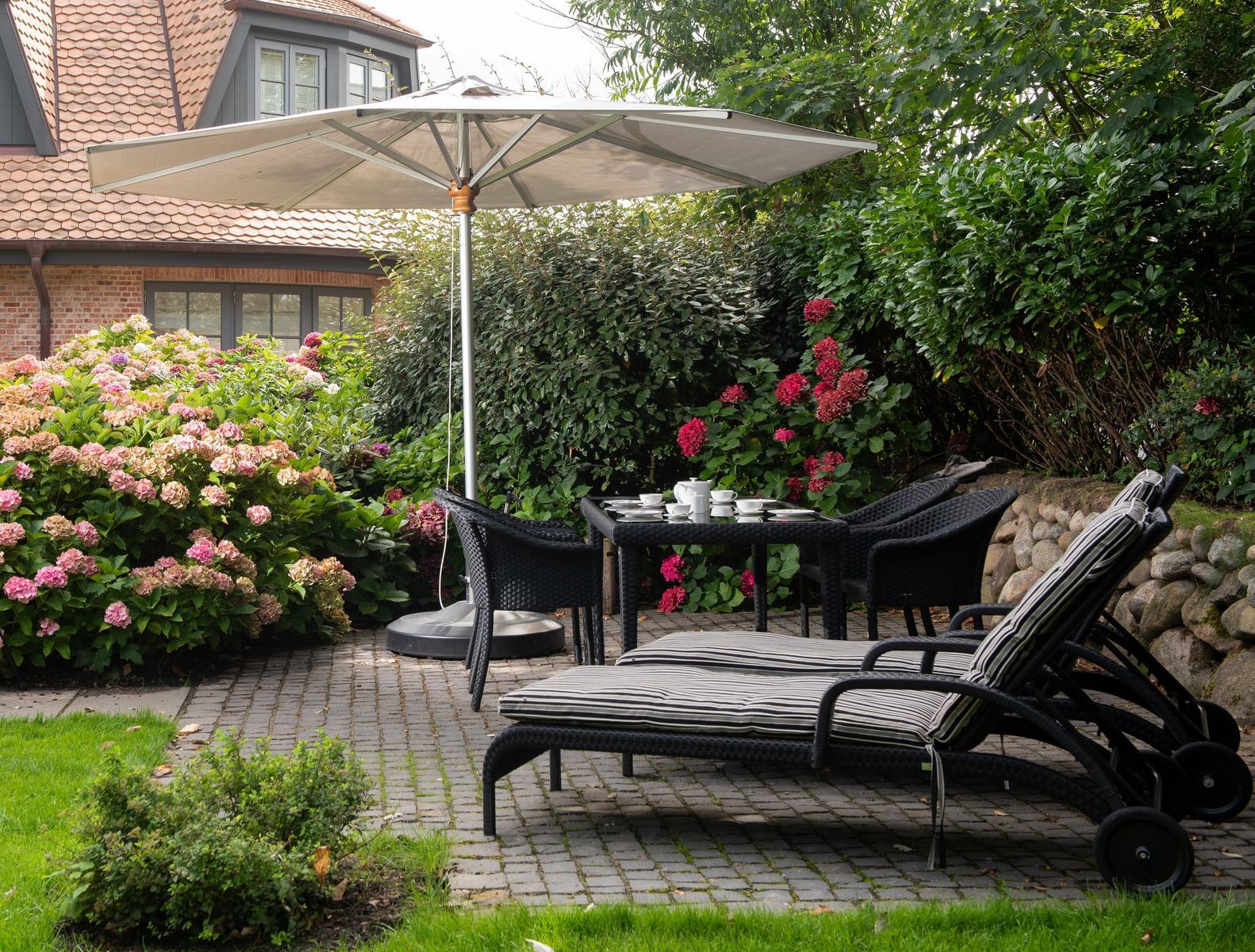 Garten mit Sonnenschirm, Tisch, Stühlen und Liegestühlen. Blumenbeete und Steinmauer umgeben den Platz.