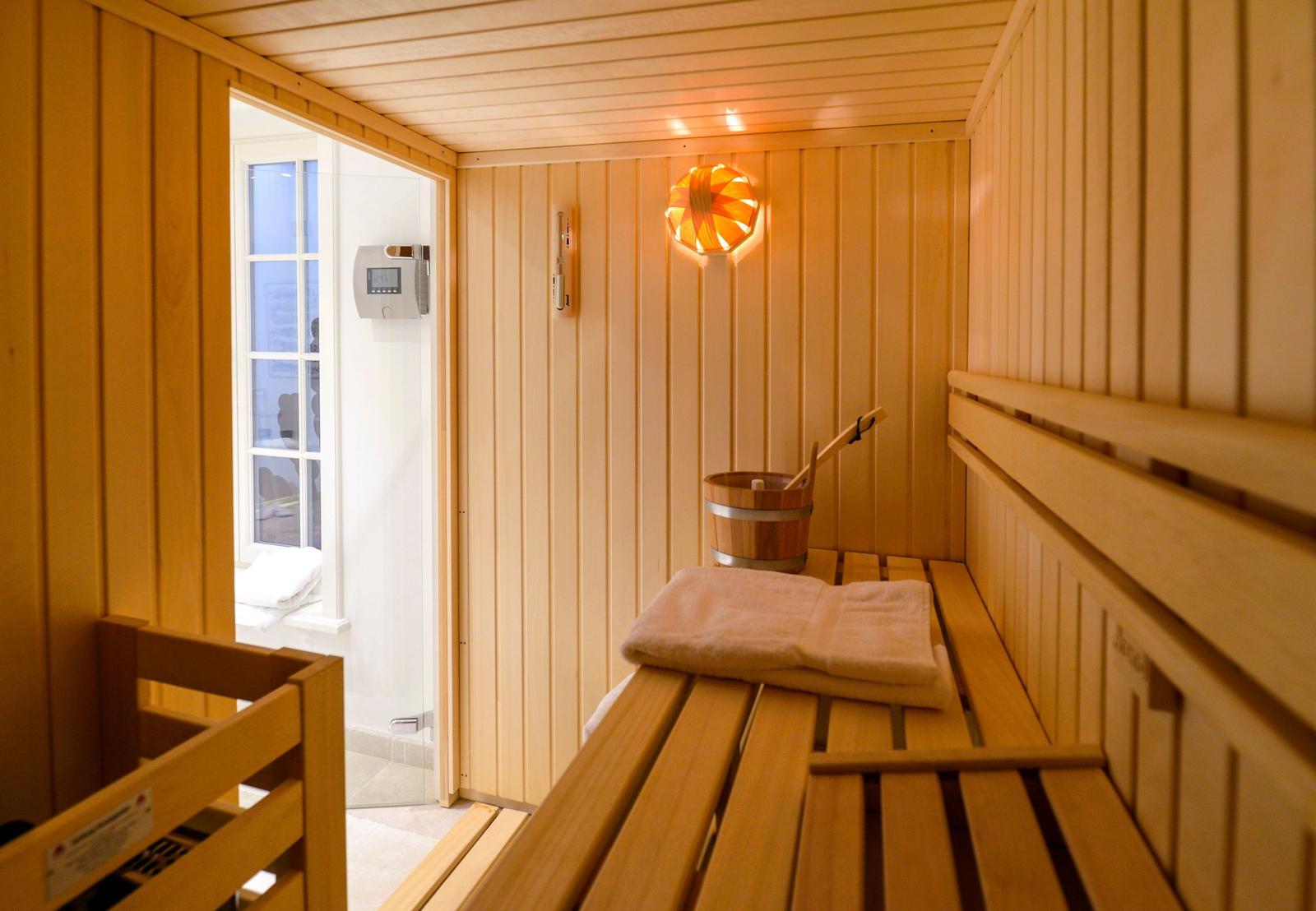 Sauna mit Holzbänken, Holzfußboden und Holzwand. Dampfheizung und Holzkrug mit Löffel.